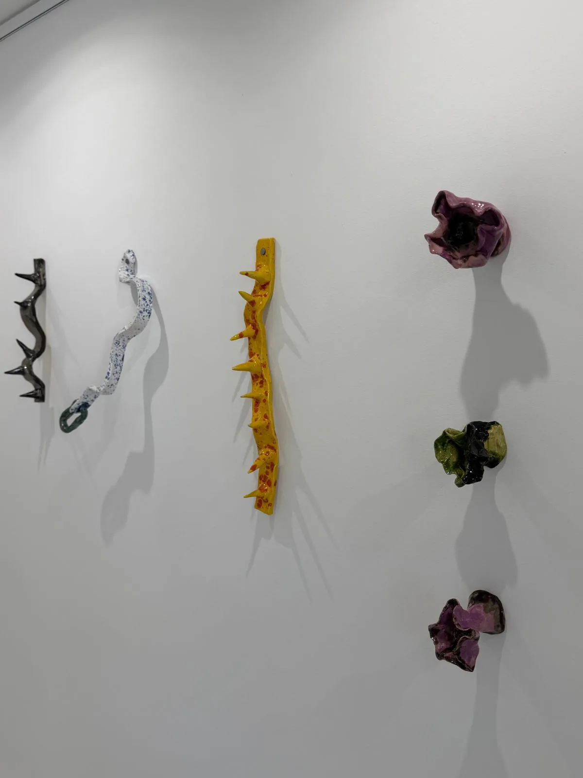 A wall of ceramics: a black thorned branch, a speckled white ribbon ending in a green ring, a yellow thorned band, and three flowers