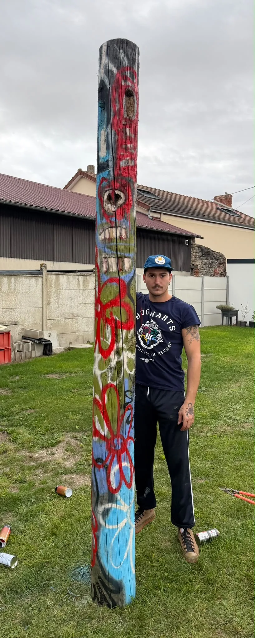 Gautier Hardy standing beside Totem (visage) in the studio garden: a tall dead trunk painted with a face, red flowers and blue spray paint